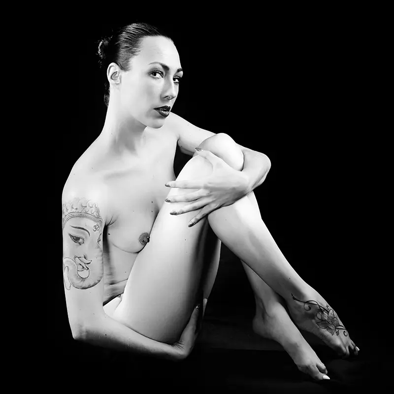 Naked woman sitting holding her legs in to her body looking at the camera with a questioning look